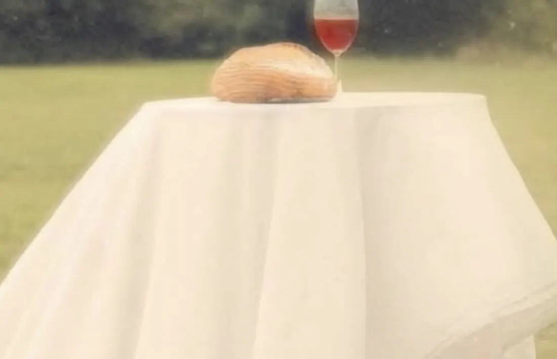 Mesa coberta com toalha branca ao ar livre, com um pão e um cálice de vinho