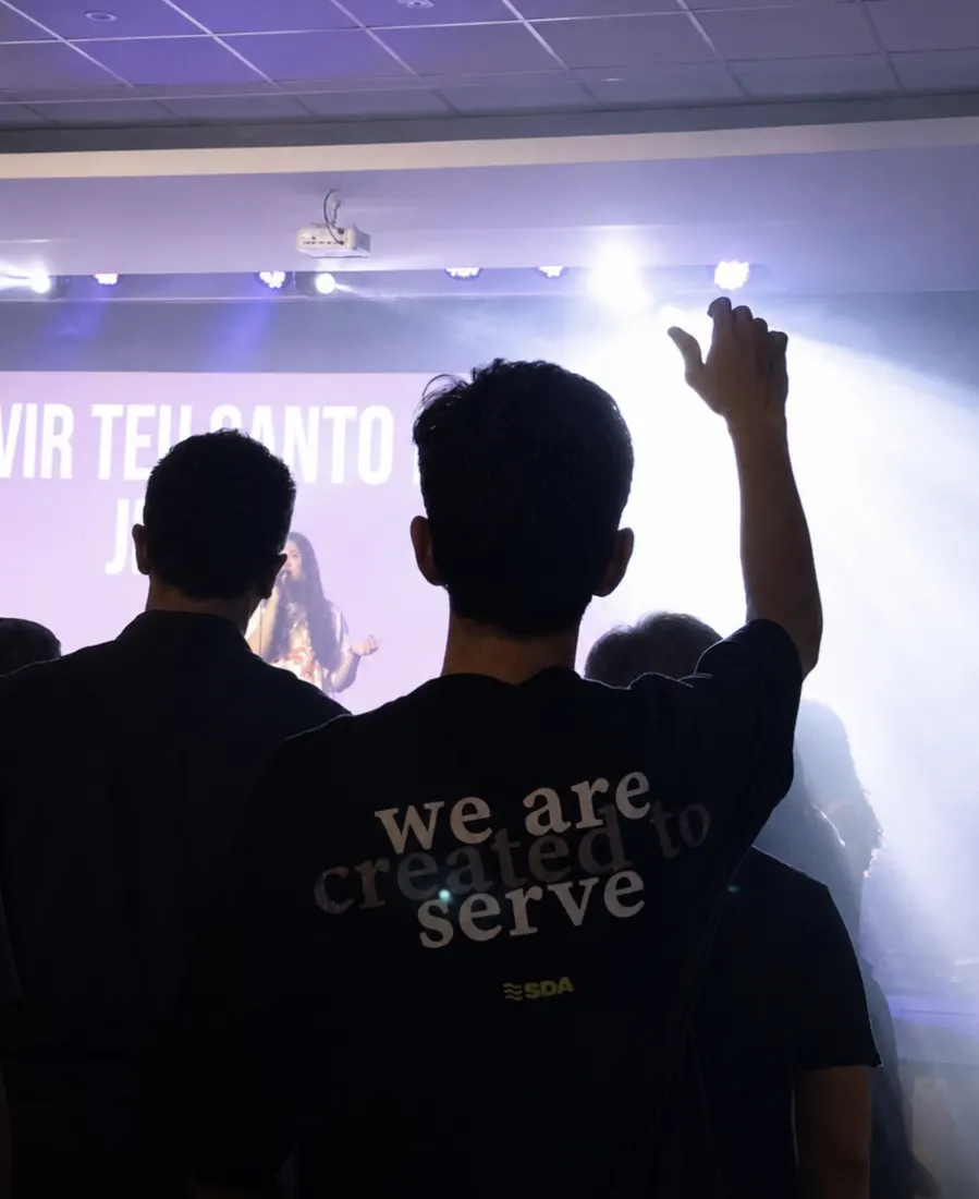 Jovem de costas, com camiseta escrita 'we are created to serve', levantando a mão no louvor